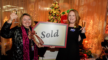 Two women holding "sold" sign in front of tree
